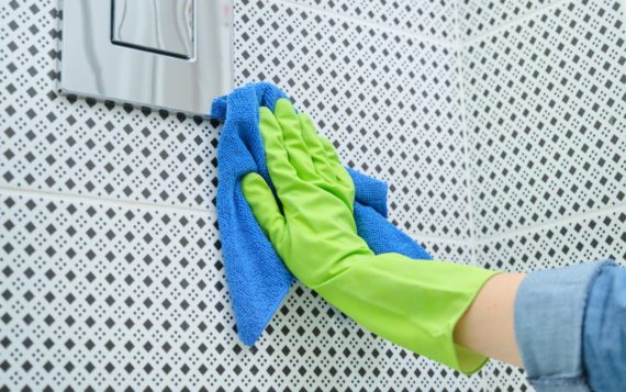 Woman in gloves with rag doing cleaning in bathroom, cleaning and polishing chrome toilet button on tiled wall